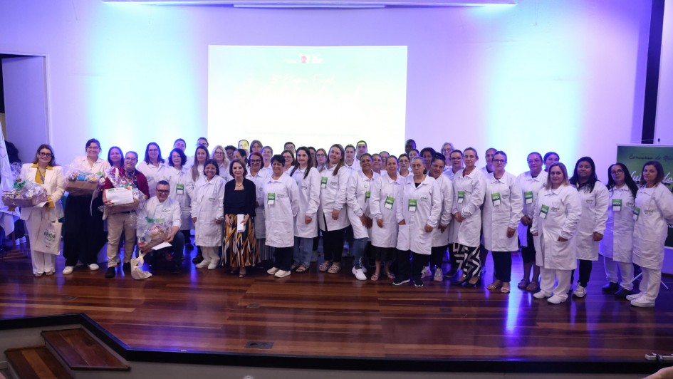 Um grande grupo de pessoas está reunido em um auditório, posicionado sobre um palco de madeira. A maioria veste jalecos brancos e crachás verdes. O grupo ocupa quase toda a largura do palco e está organizado em várias fileiras, olhando para a câmera. Em frente ao grupo há uma mulher vestindo roupas escuras e uma saia estampada, posicionada ao centro. Ao fundo, um telão iluminado em azul exibe informações sobre o evento, embora o texto não seja totalmente legível. A sala tem luzes frias que criam um brilho azul nas paredes.