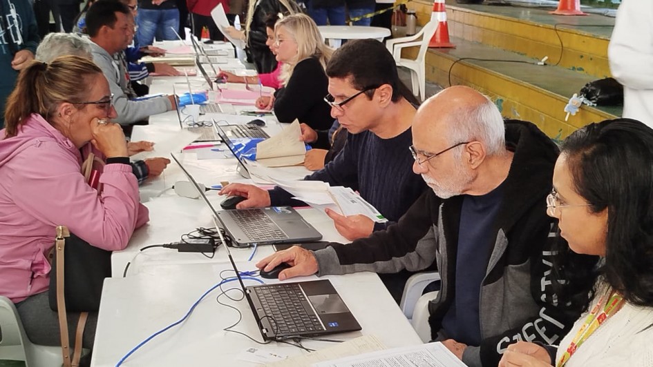 Várias pessoas estão sentadas frente a frente em mesas compridas cobertas por toalhas brancas. Em cada mesa há notebooks, documentos e canetas. Os atendentes, posicionados de um lado das mesas, digitam ou analisam papéis enquanto conversam com as pessoas atendidas. A imagem mostra um ambiente amplo, semelhante a um ginásio, com piso verde e algumas faixas de isolamento e cones laranja ao fundo. As pessoas utilizam roupas casuais e o local está movimentado, com várias fileiras de atendimento.