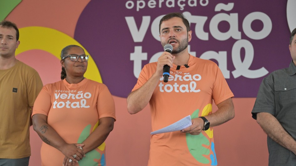 Uma foto em close-up, capturada em um evento interno, mostra um grupo de quatro pessoas paradas em frente a uma parede de fundo colorida que promove a "operação verão total".O foco principal está em um homem de pele clara no centro-direita, que está falando em um microfone que segura com a mão direita. Ele veste uma camiseta laranja promocional com o logotipo "operação verão total" em letras brancas. Ele segura um pedaço de papel dobrado na mão esquerda e usa um relógio de pulso preto.Ao lado dele, no centro-esquerda, está uma mulher de pele escura com cabelo curto e óculos. Ela também veste a mesma camiseta laranja promocional e tem os braços cruzados na frente.Outros dois homens aparecem parcialmente:À esquerda, um homem de pele clara com uma camiseta escura.À extrema direita, um homem de pele clara em uma camisa cinza-escura.O fundo é um painel de parede com as palavras "operação verão total" escritas em branco em grandes faixas horizontais de cores laranja, roxo e verde-água. A faixa roxa, mais proeminente, contém a palavra "verão" e o til ($\tilde{}$) alongado.