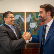 Foto em plano americano de Davi e Leite de lado. Eles estão um de frente para o outro e se dão as mãos, sorrindo.