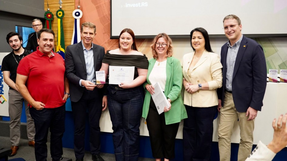 Oito pessoas estão alinhadas em frente a um painel de apresentação em uma sala de eventos. No centro, uma jovem segura um certificado, enquanto a pessoa ao lado segura um troféu com o texto parcialmente visível “Campeões de Futuro”. As demais pessoas ao redor sorriem para a câmera. Ao fundo, há bandeiras posicionadas atrás do grupo, incluindo a bandeira do Brasil. À direita, sobre uma mesa, há outros troféus iguais alinhados. O ambiente é iluminado, com parede de fundo em tons claros e uma tela de projeção exibindo parte do nome do programa.