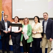Oito pessoas estão alinhadas em frente a um painel de apresentação em uma sala de eventos. No centro, uma jovem segura um certificado, enquanto a pessoa ao lado segura um troféu com o texto parcialmente visível “Campeões de Futuro”. As demais pessoas ao redor sorriem para a câmera. Ao fundo, há bandeiras posicionadas atrás do grupo, incluindo a bandeira do Brasil. À direita, sobre uma mesa, há outros troféus iguais alinhados. O ambiente é iluminado, com parede de fundo em tons claros e uma tela de projeção exibindo parte do nome do programa.