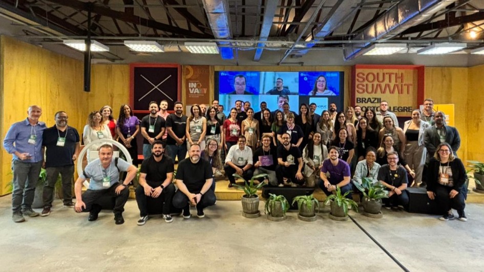 Foto posada de mais de 50 pessoas em pé ou agachadas em ambiente fechado, com um telão ao fundo.