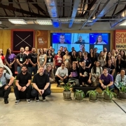 Foto posada de mais de 50 pessoas em pé ou agachadas em ambiente fechado, com um telão ao fundo.