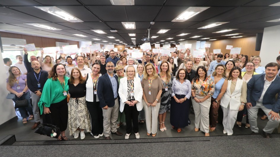 Descrição da Imagem para Acessibilidade
Uma foto de plano aberto tirada em um auditório interno mostra um grande grupo de pessoas reunidas para uma cerimônia ou evento de encerramento, provavelmente relacionado ao "Qualifica Vigilância RS 2025".

Pessoas:
Composição: O grupo é composto por uma grande quantidade de homens e mulheres, de diversas idades, alinhados em várias filas, ocupando a largura do ambiente. Há uma mistura de vestimentas casuais e sociais.

Ação Principal: Quase todas as pessoas no fundo e nas laterais estão segurando certificados ou folhas de papel branco acima da cabeça com as duas mãos, em um gesto de celebração ou conclusão de um curso.

Foco Central: Na primeira fila, no centro, há um grupo de cerca de 10 a 12 pessoas que parecem ser líderes ou organizadores. Entre elas, uma mulher de cabelos loiros e um casaco branco e preto se destaca no centro. Outras mulheres na frente vestem roupas coloridas e saias longas.

Cenário:
Teto: O teto é baixo e forrado com diversas luminárias retangulares de LED, que iluminam bem o ambiente.

Paredes: As paredes laterais são neutras, com janelas no lado esquerdo que deixam entrar alguma luz natural.

Chão: O chão é revestido com carpete cinza escuro.

Estrutura: O grupo está em pé em uma pequena elevação ou estrado forrado, indicando que estão em uma área de palco ou de apresentação.