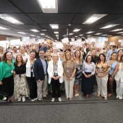 Descrição da Imagem para Acessibilidade
Uma foto de plano aberto tirada em um auditório interno mostra um grande grupo de pessoas reunidas para uma cerimônia ou evento de encerramento, provavelmente relacionado ao "Qualifica Vigilância RS 2025".

Pessoas:
Composição: O grupo é composto por uma grande quantidade de homens e mulheres, de diversas idades, alinhados em várias filas, ocupando a largura do ambiente. Há uma mistura de vestimentas casuais e sociais.

Ação Principal: Quase todas as pessoas no fundo e nas laterais estão segurando certificados ou folhas de papel branco acima da cabeça com as duas mãos, em um gesto de celebração ou conclusão de um curso.

Foco Central: Na primeira fila, no centro, há um grupo de cerca de 10 a 12 pessoas que parecem ser líderes ou organizadores. Entre elas, uma mulher de cabelos loiros e um casaco branco e preto se destaca no centro. Outras mulheres na frente vestem roupas coloridas e saias longas.

Cenário:
Teto: O teto é baixo e forrado com diversas luminárias retangulares de LED, que iluminam bem o ambiente.

Paredes: As paredes laterais são neutras, com janelas no lado esquerdo que deixam entrar alguma luz natural.

Chão: O chão é revestido com carpete cinza escuro.

Estrutura: O grupo está em pé em uma pequena elevação ou estrado forrado, indicando que estão em uma área de palco ou de apresentação.