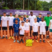 Um grupo de aproximadamente vinte pessoas, incluindo crianças, adolescentes e adultos, está reunido em uma quadra de saibro de tênis. Todos aparecem alinhados em duas fileiras diante da rede. A maior parte veste camisetas esportivas brancas ou azuis, e alguns usam bonés. No centro, à frente, há uma pilha organizada de bolas de tênis amarelas. Ao fundo, um banner roxo com texto referente ao programa “Pró-Esporte” está preso à grade de proteção da quadra. Árvores verdes e uma rua com um carro aparecem atrás da grade.