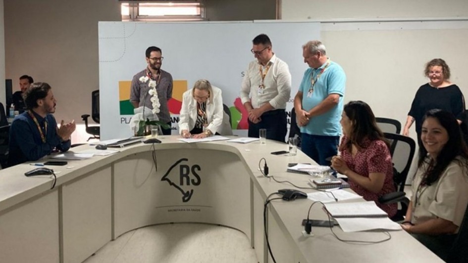 A foto mostra uma reunião em uma sala institucional, com uma mesa em formato semicircular. No centro da mesa, uma mulher inclina-se para frente enquanto assina um documento. Atrás dela, três homens estão de pé observando a assinatura. Ao lado direito da imagem, duas mulheres estão sentadas, uma delas sorrindo e a outra com um caderno aberto à sua frente. À esquerda, um homem sentado aplaude. Ao fundo, um painel exibe elementos gráficos, incluindo formas nas cores verde, amarelo e vermelho, além do texto parcialmente visível do programa estadual. O logotipo estilizado do mapa do Rio Grande do Sul com as iniciais “RS” aparece na frente da mesa.