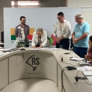 A foto mostra uma reunião em uma sala institucional, com uma mesa em formato semicircular. No centro da mesa, uma mulher inclina-se para frente enquanto assina um documento. Atrás dela, três homens estão de pé observando a assinatura. Ao lado direito da imagem, duas mulheres estão sentadas, uma delas sorrindo e a outra com um caderno aberto à sua frente. À esquerda, um homem sentado aplaude. Ao fundo, um painel exibe elementos gráficos, incluindo formas nas cores verde, amarelo e vermelho, além do texto parcialmente visível do programa estadual. O logotipo estilizado do mapa do Rio Grande do Sul com as iniciais “RS” aparece na frente da mesa.