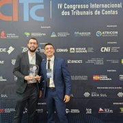 Foto mostra dois homens posando para foto, em frente a um painel de fundo preto com diversas logomarcas e o símbolo do evento. Um deles está com um troféu de acrílico.