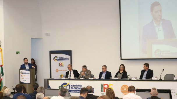 Uma foto colorida e em plano geral registra uma mesa de debate durante o "XII F&oacute;rum Econ&ocirc;mico Ga&uacute;cho", conforme indicado pelos logotipos e pain&eacute;is no ambiente.

No centro da imagem, um palestrante, um homem branco com barba e terno azul-marinho, est&aacute; de p&eacute; em um p&uacute;lpito &agrave; esquerda. Ele gesticula enquanto fala para uma plateia sentada na parte inferior da imagem. No fundo e &agrave; direita, um tel&atilde;o reproduz o close-up do palestrante.

&Agrave; direita do palestrante, sentados &agrave; mesa, est&atilde;o cinco debatedores (quatro homens e uma mulher) atentos ao discurso. No painel &agrave; frente da mesa, l&ecirc;-se "XII F&Oacute;RUM ECON&Ocirc;MICO GA&Uacute;CHO - DESAFIOS PARA O FUTURO".

A sala tem paredes brancas, e a plateia &eacute; composta por pessoas de costas, majoritariamente em trajes sociais.