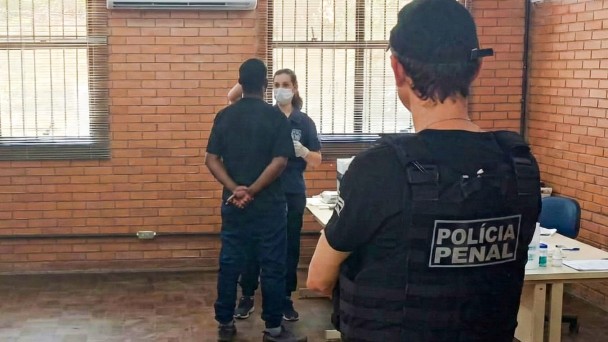 A foto mostra uma sala com paredes de tijolos aparentes e janelas amplas com persianas. No centro, uma pessoa em p&eacute;, de costas para a c&acirc;mera, est&aacute; com as m&atilde;os cruzadas atr&aacute;s do corpo enquanto conversa com uma profissional de sa&uacute;de que usa m&aacute;scara e luvas, posicionada diante de uma mesa com materiais de coleta. &Agrave; direita, um agente penal aparece em primeiro plano, tamb&eacute;m de costas, vestindo colete t&aacute;tico com a inscri&ccedil;&atilde;o &ldquo;POL&Iacute;CIA PENAL&rdquo;. No canto da sala h&aacute; uma cadeira azul e alguns itens sobre a mesa. O ambiente &eacute; iluminado pela luz natural que entra pelas janelas.