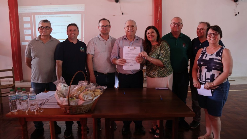 Oito pessoas estão posicionadas atrás de uma mesa de madeira. No centro, um homem segura um documento impresso, com uma mulher ao lado apoiando o papel com uma das mãos. Os demais estão alinhados, olhando para a câmera. Sobre a mesa, há uma cesta com pães e biscoitos embalados, além de garrafas de água, copos, documentos e canetas. Ao fundo, uma projeção é exibida na parede branca, com textos parcialmente visíveis.