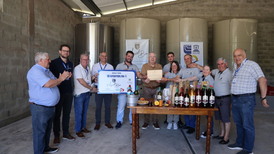 Uma fotografia em plano geral registra um grupo de treze pessoas, majoritariamente homens, posando em torno de uma mesa com garrafas de vinho, em um ambiente interno que se assemelha a uma vinícola ou agroindústria. O grupo celebra o recebimento de uma certificação ou premiação.

Pessoas e Posições
O grupo é composto por treze pessoas (onze homens e duas mulheres) vestindo, em sua maioria, trajes casuais.

Centro-Esquerda: Um pequeno grupo de homens está à esquerda, com um deles (o segundo da esquerda) segurando um certificado ou placa azul e branco que parece ser uma premiação. Outros homens próximos a ele estão batendo palmas.

Centro-Direita: Um homem mais velho, no centro, segura um certificado em papel pardo (cor de papelão) com as duas mãos, posando orgulhosamente. Uma mulher e um homem mais jovens estão atrás dele.

Extremidades: O grupo se estende horizontalmente, com a maioria das pessoas sorrindo e olhando para a câmera.

Cenário e Decoração
Ambiente Industrial: O local parece ser o interior de um armazém ou área de processamento. O chão é de concreto, as paredes são de tijolos à vista (cimento queimado/alvenaria bruta), e o teto é metálico com telhas de zinco.

Tanques: No fundo, visíveis à esquerda, há grandes tanques cilíndricos, provavelmente de aço inoxidável e fibra (ou concreto revestido) utilizados no processo de fermentação ou armazenamento de vinho.

Mesa de Exposição: No centro, há uma rústica mesa de madeira, sobre a qual estão dispostas diversas garrafas de vinho, frutas (como pêssegos e laranjas) e alguns petiscos ou alimentos.

Textos Visíveis (em Banners e Certificados)
Um banner suspenso no fundo, à esquerda, apresenta o texto em destaque: "Natureza do Bio" e o logotipo da EMATER-RS aparece em outro banner menor e também no certificado azul/branco, que diz "AQUI TEM EMATER/RS".

A foto transmite uma sensação de sucesso, parceria e celebração da produção agroindustrial.