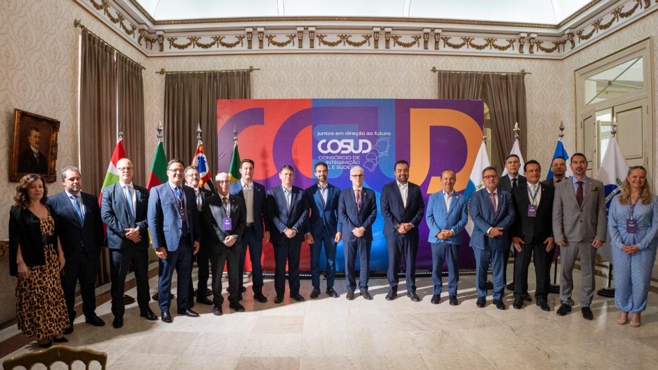 Grupo de autoridades posa em frente ao painel colorido com tema de integração regional. Ambiente solene com bandeiras e decoração clássica.