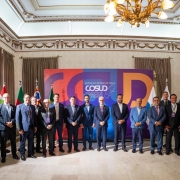 Grupo de autoridades posa em frente ao painel colorido com tema de integração regional. Ambiente solene com bandeiras e decoração clássica.