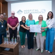 A imagem é uma fotografia de grupo que documenta a assinatura de um convênio para a compra de um mamógrafo pelo Hospital Sapiranga, no Rio Grande do Sul.

Elementos da Cena:
Grupo de Pessoas: Seis pessoas estão alinhadas no centro da imagem, sorrindo e olhando para a câmera.

No centro, duas mulheres estão segurando um documento (o convênio assinado). A mulher da esquerda veste uma blusa turquesa, e a mulher da direita veste um blazer verde-claro.

As outras quatro pessoas (dois homens nas pontas esquerda e centro-esquerda, e duas mulheres nas pontas direita e centro-direita) estão posicionadas ao lado delas, acompanhando o momento.

A maioria das pessoas usa crachás no pescoço.

Fundo (Parede/Painel):

A parede de fundo é clara, com um grande painel digital exibindo o logotipo e nome do "HOSPITAL SAPIRANGA".

À direita do logotipo do hospital, há um selo ou pequeno logotipo da "Secretaria da Saúde - OMS" ou similar.

No canto esquerdo, há um painel de madeira vertical com detalhes de textura, e o logotipo do hospital é repetido em uma placa menor ao lado.

Mobiliário (Primeiro Plano):

No canto inferior esquerdo, há uma pequena mesa de centro de madeira escura com seis garrafas de água mineral dispostas em duas filas.

Cadeiras claras e modernas são visíveis na lateral esquerda.

Conclusão Visual:
A fotografia celebra um evento institucional importante para o Hospital Sapiranga, possivelmente o anúncio ou a formalização de recursos para a aquisição de um mamógrafo, com a presença de autoridades e representantes.