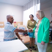 A imagem mostra um quarto hospitalar com um homem idoso sentado à beira de uma cama, estendendo a mão para cumprimentar uma mulher em pé. A cama está posicionada à esquerda, com lençóis brancos e um painel de cabeceira contendo interruptores e tomadas. Atrás do homem há uma mulher em pé, observando a cena. À direita, a secretária Arita cumprimenta o paciente. Ela veste blazer verde claro e crachá. Ao fundo, há outro homem em pé ao lado de uma segunda cama. Cortinas brancas dividem os espaços do quarto.