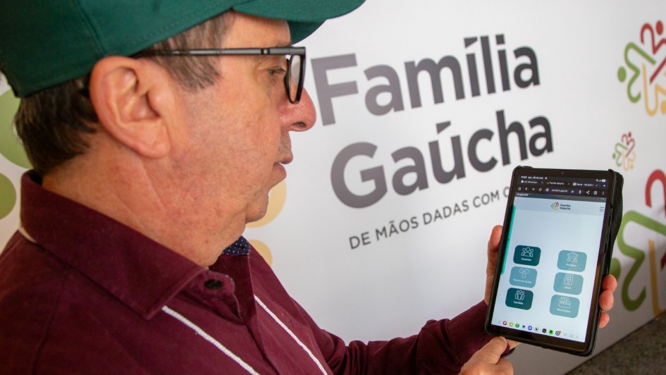 A imagem é um close-up de um homem adulto segurando e interagindo com um tablet digital, com um painel de fundo que anuncia o "Programa Família Gaúcha".

Elementos da Cena:
Homem:

Veste uma camisa polo ou social de cor vinho/bordô e um boné verde escuro.

Usa óculos de grau de armação preta e está olhando fixamente para a tela do tablet.

Sua mão direita está segurando e interagindo com a tela do dispositivo.

Tablet (Plano Central):

O tablet exibe um aplicativo ou página web com o cabeçalho "Família Gaúcha" e um menu de opções em ícones e caixas, sugerindo uma plataforma de cadastro ou acompanhamento de programas sociais.

Fundo (Painel):

A imagem tem como fundo um grande painel branco com o logotipo e nome do programa, que é "Família Gaúcha" em letras grandes, em tons de cinza.

Abaixo do nome, está o slogan "DE MÃOS DADAS COM O" (o restante do slogan é cortado).

O painel também tem símbolos gráficos coloridos e estilizados (vermelho, verde, laranja) no canto superior direito.

Conclusão Visual:
A foto ilustra uma pessoa utilizando a plataforma digital do Programa Família Gaúcha, provavelmente no contexto do processo de seleção ou acompanhamento das famílias beneficiadas, conforme o título do arquivo ("Programa Família Gaúcha finaliza seleção de 10.048 famílias").