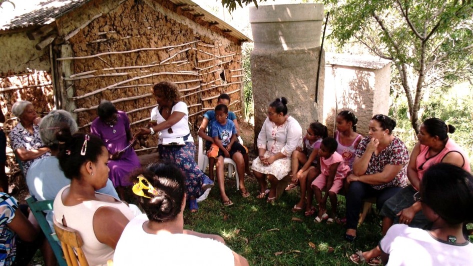 A foto mostra um grupo de pessoas reunidas em círculo ao ar livre, sentadas em cadeiras e bancos sobre a grama, à sombra de árvores. São principalmente mulheres, além de algumas crianças e duas pessoas idosas. No centro do círculo, uma mulher interage com uma senhora sentada ao seu lado. Ao fundo, há uma casa de barro com estrutura de madeira exposta e um pequeno anexo de concreto. A vegetação ao redor é densa, com árvores projetando sombra sobre o grupo.