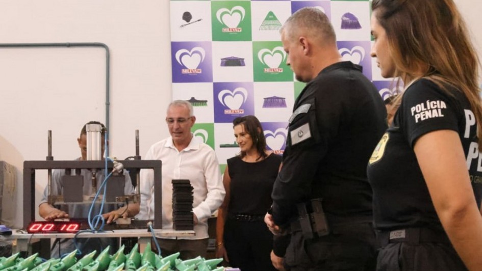 A imagem é uma fotografia em ambiente fechado (industrial) que mostra uma nova frente de trabalho para apenados na Penitenciária de Porto Alegre, em parceria com uma indústria gaúcha (logotipo Milaza ao fundo).

Elementos da Cena:
Primeiro Plano (Trabalho/Produtos):

Uma longa fileira de peças plásticas verdes (possivelmente cabos de vassouras ou outros utensílios, com formato triangular na ponta) está disposta na diagonal, pronta para serem processadas ou embaladas.

Plano Central (Ação e Pessoas):

Operário Apenado (Esquerda): Um homem (presumivelmente um apenado) está sentado atrás de uma máquina industrial (prensa ou montadora). Ele está focado na operação da máquina, processando pequenos pedaços pretos (possivelmente as peças que serão acopladas aos cabos verdes).

Autoridades/Visitantes (Centro e Direita):

Homem de Camisa Branca (Centro-Esquerda): Um homem de meia-idade, vestindo camisa social branca, está observando o trabalho na máquina.

Mulher de Preto (Centro): Uma mulher, vestindo roupa preta sem mangas, observa o processo de trabalho ao lado.

Policiais Penais (Direita): Um homem e uma mulher, ambos vestindo uniformes pretos (o homem com insígnia no ombro e a mulher com o texto "POLÍCIA PENAL" na camiseta), estão de costas para a câmera ou de perfil, supervisionando a área de trabalho.

Fundo (Institucional):

A parede de fundo é branca e exibe um painel com o logotipo da empresa parceira, "MILAZA". O logotipo é composto por corações sobrepostos em tons de verde e roxo.

Conclusão Visual:
A foto documenta a inauguração ou visita a uma oficina de trabalho dentro da Penitenciária de Porto Alegre, evidenciando a parceria entre o governo (Polícia Penal) e uma empresa privada (Milaza) na ressocialização dos apenados através do trabalho industrial.