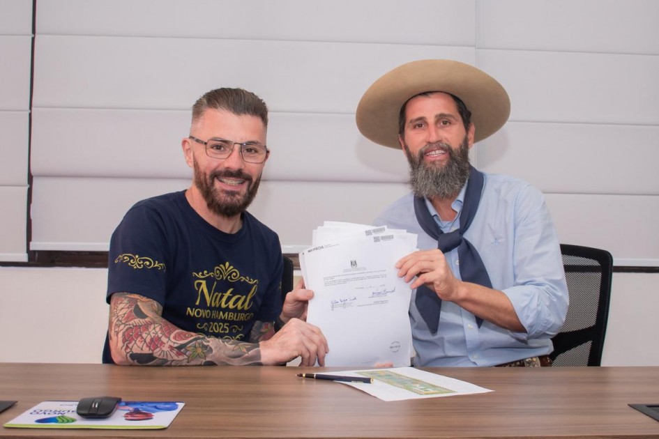 Foto mostra o Prefeito de Novo Hamburgo, Gustavo Finck, de camiseta azul escura à esquerda, e o secretário de Esporte e Lazer, Juliano Franczak, o Gaúcho da Geral, de camisa azul clara, lenço gaúcho azul escuro e chapéu bege da aba larga. Eles estão sentados lado a lado, mostrando um documento em que seguram juntos. Os dois sorriem para a câmera.