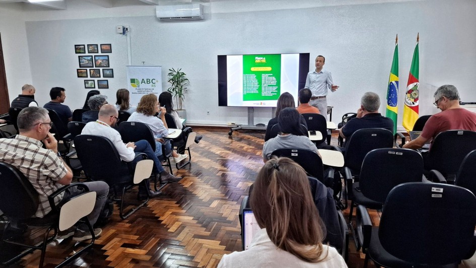 A foto mostra uma sala de reuniões ampla, com fileiras de cadeiras pretas onde várias pessoas estão sentadas voltadas para a frente. Um homem em pé, à direita da imagem, faz uma apresentação diante de uma tela grande que exibe um slide com fundo verde e textos. Ao lado da tela, estão posicionadas duas bandeiras: a do Brasil e a do Rio Grande do Sul. Na lateral esquerda há um banner com o texto “ABC+ RS Plano Estadual de Agricultura de Baixa Emissão de Carbono”. O piso é de madeira em padrão escuro, e na parede ao fundo há quadros com fotografias variadas. A sala tem iluminação clara e ar-condicionado instalado acima da tela.