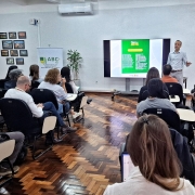 A foto mostra uma sala de reuniões ampla, com fileiras de cadeiras pretas onde várias pessoas estão sentadas voltadas para a frente. Um homem em pé, à direita da imagem, faz uma apresentação diante de uma tela grande que exibe um slide com fundo verde e textos. Ao lado da tela, estão posicionadas duas bandeiras: a do Brasil e a do Rio Grande do Sul. Na lateral esquerda há um banner com o texto “ABC+ RS Plano Estadual de Agricultura de Baixa Emissão de Carbono”. O piso é de madeira em padrão escuro, e na parede ao fundo há quadros com fotografias variadas. A sala tem iluminação clara e ar-condicionado instalado acima da tela.