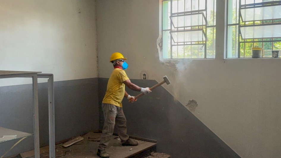 Foto de corpo inteiro de um trabalhador, usando capacete, óculos de proteção e marreta, quebrando uma estrutura na parede interna do presídio. 