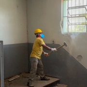 Foto de corpo inteiro de um trabalhador, usando capacete, óculos de proteção e marreta, quebrando uma estrutura na parede interna do presídio. 