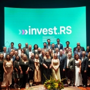 A foto mostra um grande grupo de pessoas posando para uma foto oficial em frente a um telão azul-esverdeado com o logotipo “invest.RS” e a frase “Promovendo o desenvolvimento do Rio Grande do Sul”. O grupo é composto por homens e mulheres organizados em várias fileiras. Em primeiro plano, há mesas com arranjos florais e taças com bebida. O ambiente é um auditório iluminado com luz cênica.