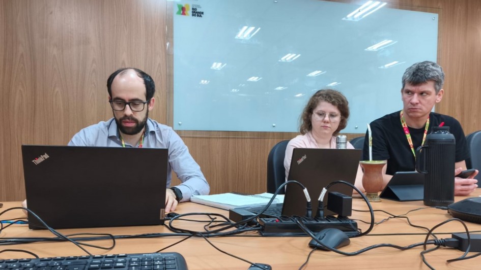 Foto de três pessoas sentadas junto a uma mesa, em frente a notebooks.
