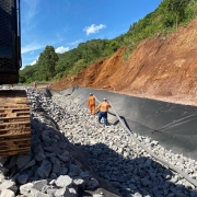 A imagem é uma fotografia em plano fechado que captura uma frente de trabalho em uma rodovia (VRS-826, entre Feliz e Alto Feliz), focada na contenção de encostas e recuperação da estrada.

Elementos da Cena:
Primeiro Plano (Trabalho):

O lado esquerdo é dominado pela parte inferior de uma máquina pesada (provavelmente uma escavadeira ou trator), com sua esteira (rastros) de cor marrom-avermelhada e ferrugem visível.

O solo na área de trabalho está coberto por uma grande quantidade de pedras soltas (rachão) de cor cinza-clara, que formam o leito da recuperação.

Plano Central (Obras):

Uma manta geotêxtil preta está estendida sobre o terreno, cobrindo o talude (a encosta) e parte da área onde as pedras estão sendo colocadas. Essa manta serve para contenção e drenagem.

Dois operários estão presentes:

Um está de costas (à esquerda), vestindo um macacão de trabalho laranja e um capacete branco.

O outro (à direita) veste uma camisa laranja e calça azul-escura, e está caminhando sobre as pedras.

Um tubo de drenagem preto está posicionado sobre as pedras, próximo à manta.

Plano de Fundo (Encosta):

À direita, a encosta do morro é alta, com terra em tom vermelho-ferrugem exposta, indicando um deslizamento ou trabalho de corte recente.

A parte superior da encosta e o fundo da imagem são cobertos por vegetação densa de cor verde (mata nativa ou arbustos).

Céu: O céu está azul e claro, com luz solar intensa, indicando um dia de tempo bom para a realização das obras.

Conclusão Visual:
A foto evidencia o trabalho em andamento de recuperação da rodovia, com foco na estabilização do talude e drenagem, após danos causados por deslizamentos ou erosão (conforme sugerido pela terra vermelha exposta na encosta).