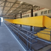 ♿ Descrição da Imagem para Acessibilidade
Esta é uma fotografia em plano médio de uma rampa de acesso para pedestres e cadeirantes, localizada em uma área coberta, possivelmente na entrada ou dentro de um prédio escolar ou público.

A rampa, no centro e à esquerda, possui piso cinza escuro e uma inclinação suave. É equipada com corrimãos duplos de metal cinza (guarda-corpos) em ambos os lados, garantindo acessibilidade e segurança. No lado direito da rampa, o corrimão é contínuo e segue a inclinação.

No centro da imagem, uma grande parede lateral, que acompanha a rampa, é pintada em um chamativo e sólido amarelo vivo. A estrutura amarela se eleva e termina em uma escada na extremidade direita.

Em primeiro plano, na base da rampa, notam-se faixas de piso tátil de alerta na cor amarela, indicando o início da inclinação.

Um pedestre (um homem vestindo camiseta azul escura e calças claras) está subindo a rampa, caminhando de costas para o observador, no terço esquerdo da imagem.

A cobertura é composta por uma estrutura metálica de treliças e telhado translúcido ou de metal claro, que permite a entrada de luz natural. O teto possui luminárias embutidas ou suspensas.

Ao fundo, à direita, a estrutura amarela se conecta a uma parede de um edifício de cor marrom (ou revestimento escuro), com diversas janelas horizontais e pequenas em sua parte superior, alinhadas em fileiras.