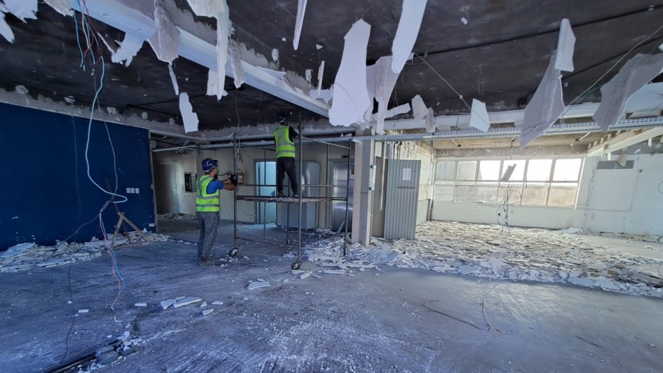 Esta é uma fotografia de um ambiente interno em processo de demolição ou reforma pesada, sob iluminação natural que entra pelas janelas ao fundo.
O ambiente geral é um salão amplo e vazio. O chão de concreto está coberto por entulho, principalmente pedaços de drywall (gesso acartonado) branco esfarelado. Fios elétricos soltos e cabos pendurados estão espalhados pelo chão e pelo teto.
O teto é escuro (preto ou cinza escuro) e mostra a estrutura original após a remoção de parte do forro. Pedaços grandes e irregulares de forro branco estão pendurados ou caindo do teto, criando uma aparência desordenada.
No centro da imagem, dois trabalhadores da construção civil estão realizando o serviço:
Um trabalhador, vestindo capacete de segurança azul e colete refletivo verde-limão, está no chão, segurando o que parece ser uma ferramenta de corte ou demolição.
O segundo trabalhador, também com capacete e colete, está sobre um andaime móvel de metal com rodízios, manipulando o teto.
O fundo da imagem é iluminado pela luz natural que entra por uma fileira de janelas horizontais e longas, que ocupam a parede direita e o final da sala. O restante das paredes internas é de concreto ou drywall, sendo que a parede do fundo é mais clara e a parede da esquerda, parcialmente visível, é pintada de um azul escuro.
A cena transmite a sensação de uma obra em andamento, com foco na remoção e desmonte das estruturas internas de um escritório ou edifício comercial.