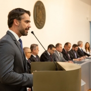 Esta é uma fotografia de um evento solene ou cerimônia formal em um ambiente interno.

Em primeiro plano e à esquerda, está o Governador Eduardo Leite, em perfil (meio-corpo), discursando em um pódio.

Ele veste um terno cinza escuro, camisa social branca e gravata azul.

Ele tem barba e cabelo escuros e está olhando ligeiramente para a frente e para a direita.

Suas mãos estão visíveis, apoiadas no pódio.

O pódio é moderno, de formato curvo e cor bege ou cáqui escura. Um microfone de pedestal preto está posicionado para captar a fala do governador.

Ao fundo, no centro, uma longa mesa cinza se estende para a direita, onde estão sentadas diversas autoridades e convidados. Eles estão vestidos com trajes formais (ternos escuros e togas pretas, sugerindo um evento de posse ou tribunal).

A maioria das pessoas sentadas são homens de meia-idade ou mais velhos, mas há algumas mulheres.

Eles estão olhando na direção do orador.

A decoração da sala é minimalista:

As paredes são lisas e de cor bege ou creme clara.

No centro da parede de fundo, acima da linha das cabeças, há um grande emblema ou brasão redondo e escuro (marrom ou bronze), possivelmente o símbolo do Tribunal de Contas do Estado (TCE).

No canto inferior direito, uma composição floral com predominância de flores laranjas e amarelas está parcialmente visível.

A iluminação é quente e concentra-se no orador em primeiro plano, enquanto o fundo está em sombra suave.
