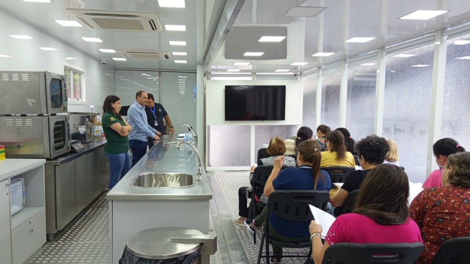 Foto do interior de uma unidade m&oacute;vel de cozinha industrial. &Agrave; esquerda, bancadas de inox com pia, equipamentos industriais, forno e micro-ondas. Tr&ecirc;s instrutores est&atilde;o de p&eacute; conversando pr&oacute;ximos &agrave; bancada. &Agrave; direita, v&aacute;rias pessoas sentadas em cadeiras pretas, de frente para uma TV grande fixada na parede frontal. O ambiente &eacute; iluminado por pain&eacute;is de LED no teto e possui janelas laterais.