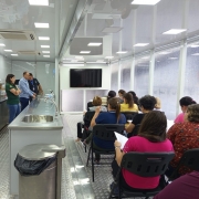 Foto do interior de uma unidade móvel de cozinha industrial. À esquerda, bancadas de inox com pia, equipamentos industriais, forno e micro-ondas. Três instrutores estão de pé conversando próximos à bancada. À direita, várias pessoas sentadas em cadeiras pretas, de frente para uma TV grande fixada na parede frontal. O ambiente é iluminado por painéis de LED no teto e possui janelas laterais.