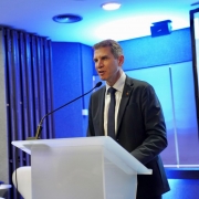 Foto de Polo falando ao púlpito, em ambiente fechado. Ao fundo há uma projeção de slide de cor azul. 