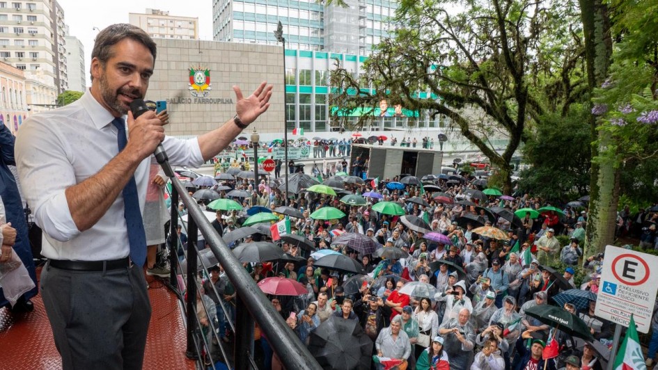 Foto de Leite falando ao microfone, em cima de um trio elétrico. Há uma grande concentração de pessoas na praça, acompanhando em capas de chuva.