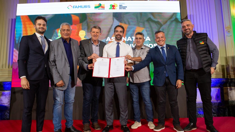 ✍️ Descrição da Imagem para Acessibilidade
Esta é uma fotografia em plano médio de um grupo de oito homens posando para a foto em um evento de assinatura de convênios, sobre um palco com fundo de mídia (tela).

O Grupo (da esquerda para a direita):

Homem 1: Jovem, veste terno azul-marinho ajustado, camisa social branca e gravata cinza-claro. Ele tem barba e cabelo escuros, e está sorrindo.

Homem 2: Mais velho, veste blazer cinza, camisa azul-escura e calças jeans. Ele tem cabelo e barba grisalhos, e está sorrindo.

Homem 3: Veste um blazer cinza-médio, camisa social e gravata escura. Ele tem cabelo grisalho-claro, e está sorrindo.

Governador Eduardo Leite (Centro): Veste camisa social branca com as mangas longas e gravata azul-escura. Calças sociais cinza escura. Ele está no centro da foto, segurando um documento dobrado (convênio) de capa vermelha e branca, exibindo-o para a câmera. Ele está sorrindo.

Homem 5: Veste camisa social clara (bege ou cinza) e calças jeans. Tem cabelo e barba escuros e está sorrindo. Ele está tocando o braço do Governador Leite.

Homem 6: Veste um blazer azul-marinho, camisa social azul-claro e gravata azul escura com um pin (broche). Ele é de pele clara, está sorrindo e também está tocando o documento que o Governador Leite segura.

Homem 7: Veste colete acolchoado preto sobre uma camisa escura, calças jeans escuras e tênis. Ele tem barba e cabelo curtos e escuros. Ele está sorrindo.

Homem 8 (extrema direita): Veste camisa social cinza escura, calças pretas e um paletó preto. Ele é o homem mais velho do grupo, tem cabelo branco e óculos. Ele está sorrindo.

O Fundo e o Palco:

Palco: Os homens estão de pé sobre um tapete ou carpete vermelho vibrante.

Fundo de Mídia: Diretamente atrás do grupo, há uma grande tela de LED retangular que exibe o logotipo do evento: "ASSINATURA DE CONVÊNIOS", além dos logotipos de FAMURS e IRG BRASIL RIO GRANDE DO SUL (aparentemente). O fundo da tela é uma imagem desfocada e colorida.

Decoração: A sala é grande e formal, com colunas e iluminação ambiente em tons de roxo e azul nas laterais, conferindo um ar solene.
