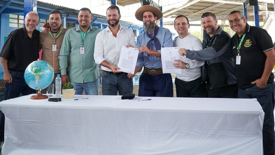 ⚽ Descrição da Imagem para Acessibilidade
Esta é uma fotografia em plano médio de um grupo de oito homens posando em um ambiente externo (possivelmente coberto, como um ginásio ou galpão), celebrando a assinatura de um documento. O evento é a assinatura de um convênio para revitalização de uma quadra esportiva em Alvorada.

O Grupo (da esquerda para a direita):

Homem 1: Sorridente, veste camisa preta e calças escuras.

Homem 2: Veste uma jaqueta marrom clara (cáqui) sobre uma camisa escura e calças escuras.

Homem 3: Veste uma camisa polo verde-clara, calças jeans e um crachá pendurado no pescoço. Ele tem barba e está sorrindo.

Homem 4 (no centro): Veste camisa social branca (com as mangas arregaçadas), calças jeans escuras. Ele tem barba e está segurando um dos documentos abertos. Ele está sorrindo.

Homem 5 (em destaque): No centro, veste trajes típicos gaúchos (pilcha): bombacha azul-escura, lenço azul no pescoço, camisa clara e um chapéu de abas largas (estilo gaúcho). Ele tem barba longa e está sorrindo, segurando o outro documento aberto.

Homem 6: Veste uma camisa branca de manga longa e calças escuras. Ele está segurando uma caneta ou apontando para um dos documentos.

Homem 7: Veste camisa polo cinza escura, calças escuras. Ele está com a mão no ombro do Homem 6, sorrindo.

Homem 8 (extrema direita): Veste uma camisa polo preta com bordados ou logotipo de um município ou organização (possivelmente Alvorada) e óculos. Ele está sorrindo.

O Fundo e os Objetos na Mesa:

Mesa: O grupo está atrás de uma mesa longa coberta por uma toalha branca, onde os documentos foram assinados.

Objetos: Sobre a mesa, no lado esquerdo, há um globo terrestre em um pedestal, garrafas de água e canetas pretas e azuis.

Fundo: O fundo é um ambiente aberto, com estruturas metálicas e paredes de madeira ou metal pintadas de azul claro e branco, indicando um galpão, ginásio ou área de lazer coberta.

A imagem é luminosa e capta a formalização de um acordo com a presença de representantes municipais, incluindo uma figura que representa a cultura local.