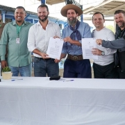 ⚽ Descrição da Imagem para Acessibilidade
Esta é uma fotografia em plano médio de um grupo de oito homens posando em um ambiente externo (possivelmente coberto, como um ginásio ou galpão), celebrando a assinatura de um documento. O evento é a assinatura de um convênio para revitalização de uma quadra esportiva em Alvorada.

O Grupo (da esquerda para a direita):

Homem 1: Sorridente, veste camisa preta e calças escuras.

Homem 2: Veste uma jaqueta marrom clara (cáqui) sobre uma camisa escura e calças escuras.

Homem 3: Veste uma camisa polo verde-clara, calças jeans e um crachá pendurado no pescoço. Ele tem barba e está sorrindo.

Homem 4 (no centro): Veste camisa social branca (com as mangas arregaçadas), calças jeans escuras. Ele tem barba e está segurando um dos documentos abertos. Ele está sorrindo.

Homem 5 (em destaque): No centro, veste trajes típicos gaúchos (pilcha): bombacha azul-escura, lenço azul no pescoço, camisa clara e um chapéu de abas largas (estilo gaúcho). Ele tem barba longa e está sorrindo, segurando o outro documento aberto.

Homem 6: Veste uma camisa branca de manga longa e calças escuras. Ele está segurando uma caneta ou apontando para um dos documentos.

Homem 7: Veste camisa polo cinza escura, calças escuras. Ele está com a mão no ombro do Homem 6, sorrindo.

Homem 8 (extrema direita): Veste uma camisa polo preta com bordados ou logotipo de um município ou organização (possivelmente Alvorada) e óculos. Ele está sorrindo.

O Fundo e os Objetos na Mesa:

Mesa: O grupo está atrás de uma mesa longa coberta por uma toalha branca, onde os documentos foram assinados.

Objetos: Sobre a mesa, no lado esquerdo, há um globo terrestre em um pedestal, garrafas de água e canetas pretas e azuis.

Fundo: O fundo é um ambiente aberto, com estruturas metálicas e paredes de madeira ou metal pintadas de azul claro e branco, indicando um galpão, ginásio ou área de lazer coberta.

A imagem é luminosa e capta a formalização de um acordo com a presença de representantes municipais, incluindo uma figura que representa a cultura local.