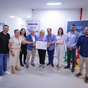 
Uma fotografia colorida captura um grupo de nove pessoas adultas, em pé, posando juntas em um ambiente interno bem iluminado, provavelmente uma instalação de saúde ou evento governamental.

Pessoas e Ação Central:
No centro da imagem, quatro pessoas estão segurando um ou mais documentos juntos, em um ato que simboliza uma assinatura, acordo ou entrega oficial.

Uma mulher de cabelos loiros curtos, vestindo uma blusa roxa, está no centro exato, segurando os papéis com ambas as mãos.

Um homem careca com camisa azul clara está à esquerda dela, também tocando os documentos.

Um homem com camisa azul escura está à direita dela, também tocando os documentos.

À extrema esquerda do grupo central, uma mulher de cabelos grisalhos, vestindo um vestido listrado bege e amarelo, sorri amplamente e aponta para os documentos ou para o grupo.

Outros Membros do Grupo:
À esquerda, um jovem de camisa preta e calças jeans está sorrindo.

Entre o jovem e a mulher de vestido listrado, estão uma mulher e um homem, ambos sorrindo.

À direita do grupo central, uma mulher de camisa rosa claro e calças brancas, e dois homens, um de camisa azul de botões e outro de camisa polo marrom, completam a formação, todos sorrindo e olhando para frente.

Fundo e Contexto:
O ambiente é de paredes claras, e há banners de propaganda ou informativos no fundo que fornecem contexto:

À esquerda, um banner roxo tem o texto "SER MULHER RS" com um símbolo.

Acima do grupo central, um cartaz azul diz "EM BREVE AQUI!" e, abaixo, o logo do SER MULHER RS se repete.

À direita, um banner azul escuro com o logo do GOVERNO DO ESTADO DO RIO GRANDE DO SUL e o texto "SECRETARIA DA SAÚDE" (indicando o local ou a natureza do evento).

O evento parece ser a celebração de uma parceria ou habilitação de um serviço de saúde.