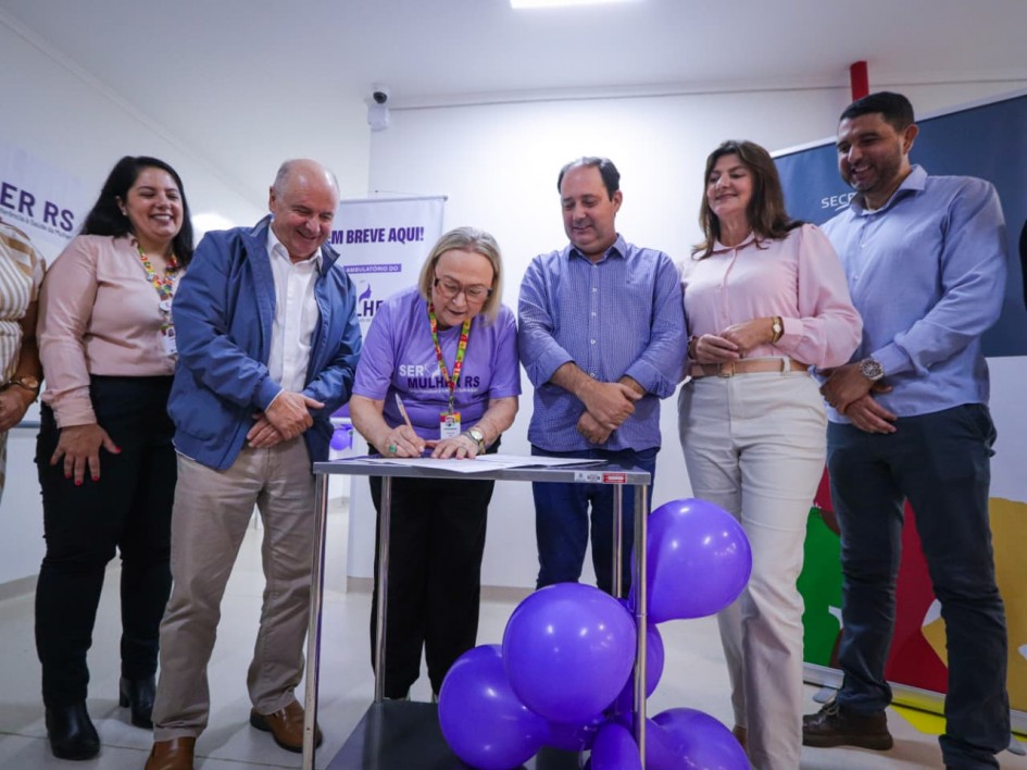 A imagem mostra um grupo de sete pessoas adultas (quatro mulheres e três homens) participando de um ato formal de assinatura, provavelmente relacionado a um programa de saúde pública em Venâncio Aires, Rio Grande do Sul, baseado no contexto do nome do arquivo (Venâncio Aires no SER Mulher RS).
Ação Central (Assinatura):
No centro, uma mulher de meia-idade, com cabelos curtos e claros e vestindo uma camiseta roxa com o logo "SER MULHER RS", está inclinada para a frente, assinando um documento que está sobre uma pequena mesa de cor escura.
Ela está concentrada na assinatura.
Pessoas Envolvidas:
À esquerda da signatária: Estão duas pessoas.
Um homem de meia-idade, vestindo uma camisa de botão azul claro e um casaco azul marinho por cima, sorri e apoia as mãos na mesa, observando a assinatura.
Uma mulher, sorrindo, com uma blusa rosa claro e uma identificação no pescoço, está atrás e à esquerda do homem.
À direita da signatária: Estão três pessoas.
Um homem de meia-idade, vestindo uma camisa de botão azul e jeans, sorri e apoia as mãos na mesa, também observando a assinatura.
Uma mulher, vestindo uma blusa rosa claro e calças brancas, sorri e está ao lado e um pouco atrás deste homem.
Um homem, vestindo uma camisa azul clara e calças escuras, sorri e está na extrema direita da formação.
Fundo e Elementos Adicionais:
O ambiente é interno, com paredes claras e boa iluminação.
No canto superior direito, um banner azul escuro exibe o logo do "GOVERNO DO ESTADO DO RIO GRANDE DO SUL" e o texto "SECRETARIA DA SAÚDE".
Acima da signatária, um cartaz pendurado diz "EM BREVE AQUI!".
Há um pequeno agrupamento de balões de festa roxos no chão, em frente ao grupo, simbolizando a cor do programa "SER MULHER RS" e o evento.