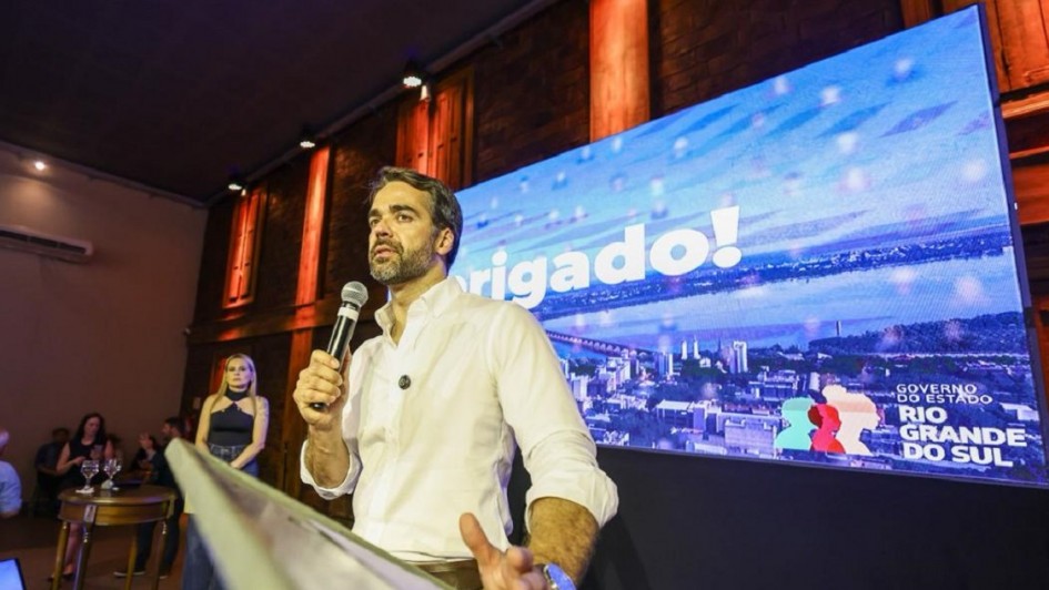 Governador Eduardo Leite está falando ao microfone em um púlpito. Atrás dele, há uma tela com uma imagem de Uruguaiana e a logo do governo do Estado.