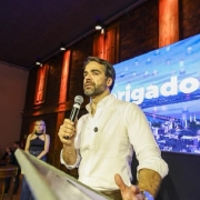 Governador Eduardo Leite está falando ao microfone em um púlpito. Atrás dele, há uma tela com uma imagem de Uruguaiana e a logo do governo do Estado.