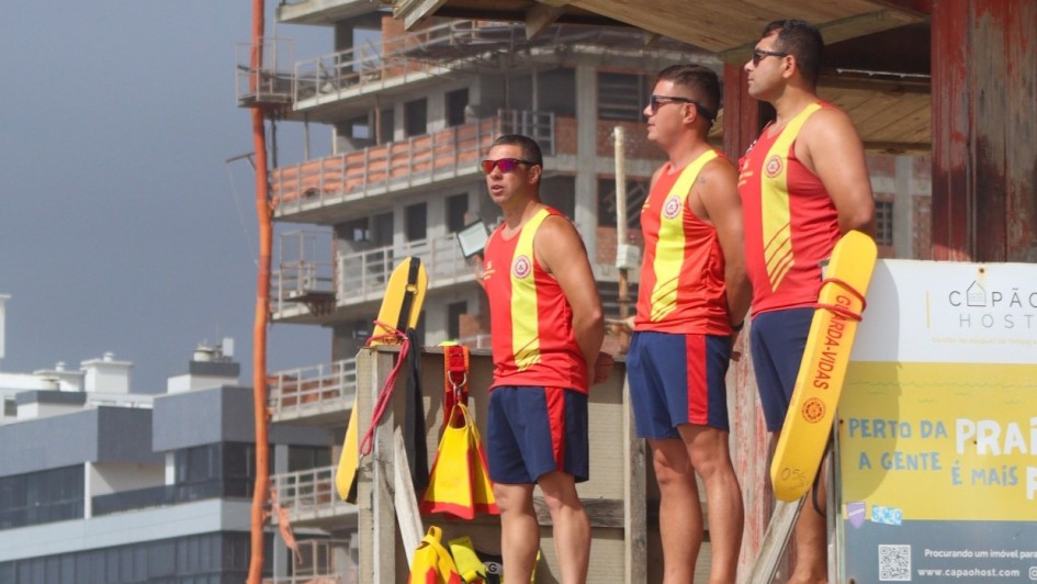 Três guarda-vidas estão posicionados em uma plataforma elevada de observação na praia. Eles vestem uniforme vermelho com faixa amarela vertical e seguram equipamentos de resgate também amarelos. Os três usam óculos escuros e observam o mar. Ao fundo, há prédios em construção e outras edificações próximas à orla.