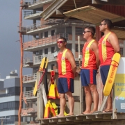 Três guarda-vidas estão posicionados em uma plataforma elevada de observação na praia. Eles vestem uniforme vermelho com faixa amarela vertical e seguram equipamentos de resgate também amarelos. Os três usam óculos escuros e observam o mar. Ao fundo, há prédios em construção e outras edificações próximas à orla.