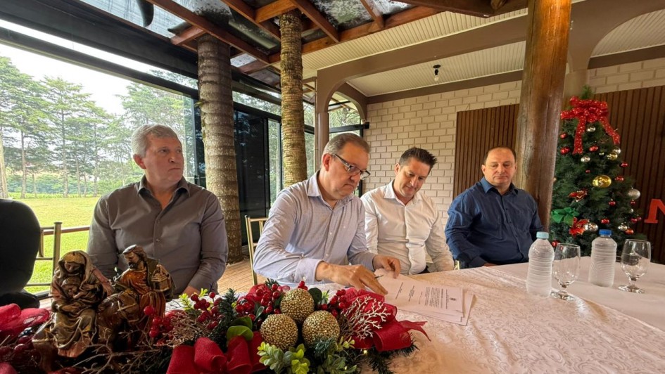 Foto mostra o secretário Edivilson Brum assinando um documento. Ele está sentado diante de uma mesa e ao seu lado, estão sentados prefeitos da região. Na mesa e no ambiente há decorações de Natal.